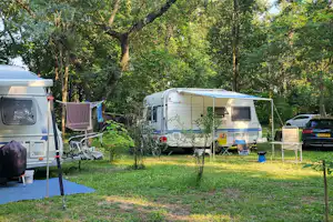 Wohnwagen und Campingbus auf einem Stellplatz im Grünen, mit Vorzelt, Wäscheleine, Stühlen und Bäumen im Hintergrund.