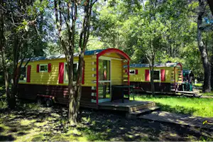 Grünes Mobilheim mit Terrasse und Eingang, umgeben von Bäumen im Campingplatz, daneben weitere Unterkünfte und Sitzbereich.