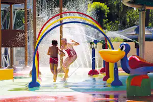 Wasserspielplatz mit bunten Rutschen und Sprühfontänen, Kinder laufen auf dem Boden im Campingbereich.