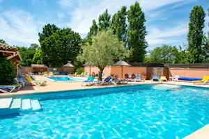 Schwimmbecken mit blauem Wasser, Liegen und Sonnenschirmen am Campingplatz, dahinter Bäume und eine Mauer.