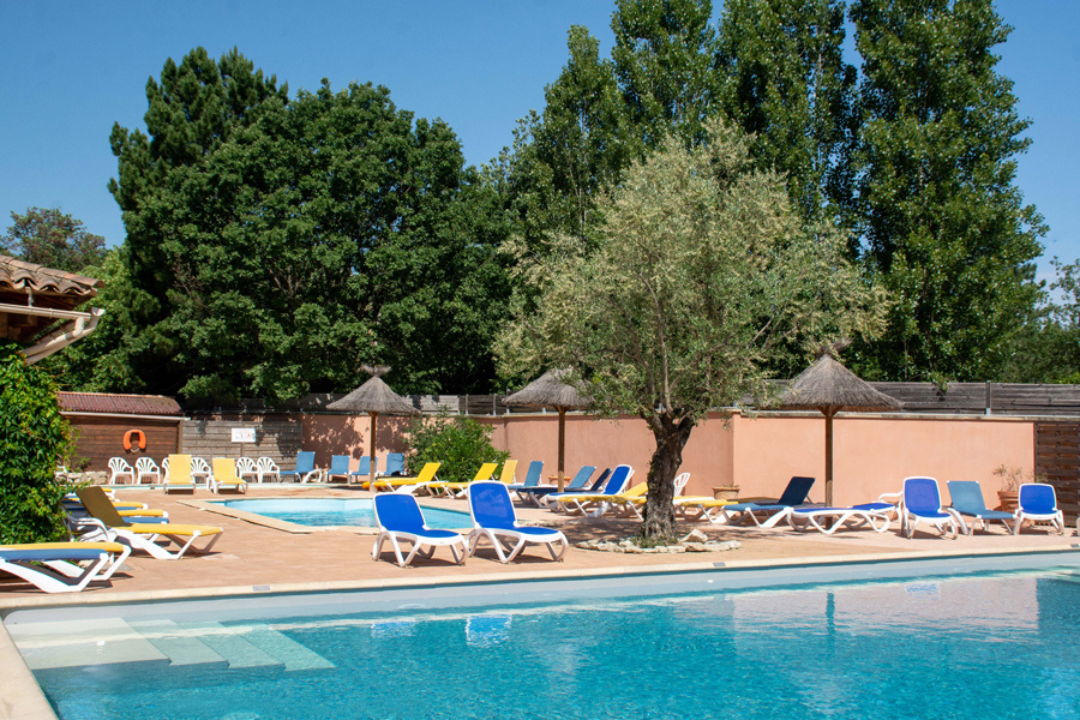 Camping-Pool mit blauen Liegen und Sonnenschirmen, umgeben von Bäumen und einer hellen Mauer.