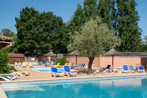 Camping-Pool mit blauen Liegen und Sonnenschirmen, umgeben von Bäumen und einer hellen Mauer.
