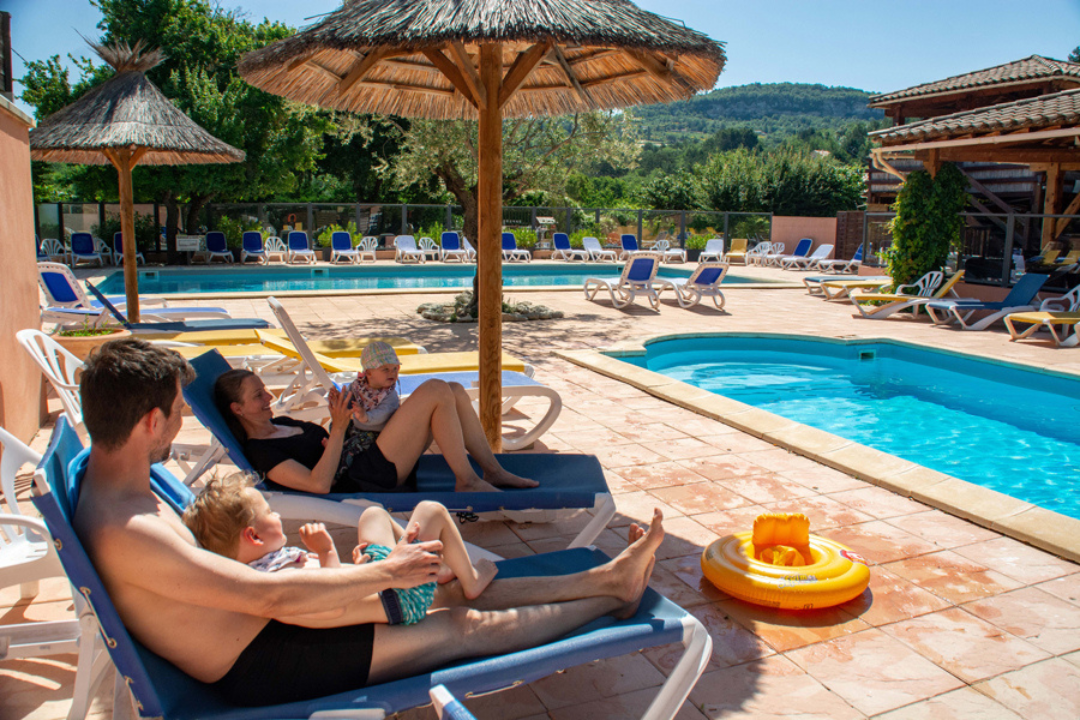 Camping-Pool mit Sonnenschirmen, Liegestühlen und gepflasterter Terrasse, im Hintergrund Bäume und Ferienhäuser.