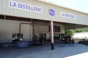 Gebaeude mit Schildern „LA DISTILLERIE“ und „LA BOUTIQUE“, Eingang mit Tor, Schuppen und Technikrohren im Hof.