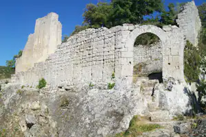 Steinruine mit Mauerbogen und hohen Mauern, umgeben von Bäumen und Felsgelände unter blauem Himmel.