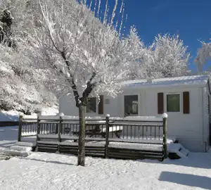 Verschneites Mobilheim mit Terrasse und Geländer, davor ein Baum mit Schnee, im Hintergrund weitere Bäume und blauer Himmel.