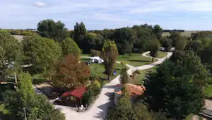 Campingplatz mit Stellplätzen, gepflasterten Wegen und kleinen Gebäuden, umgeben von Bäumen und grünen Wiesen.