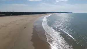 Weite Sandküste mit Strandlinie und Wellen, Blick von oben auf Meer und Uferweg im Hintergrund.