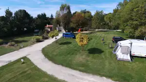 Campingplatz mit Rasenfläche, Kiesweg, Zelt und mehreren Autos, im Hintergrund Gebäude und Spielplatz im Grünen.