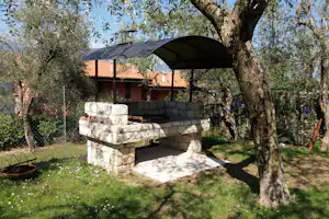 Überdachter Grillplatz mit Steinmauer, Bank und Tisch im Garten, umgeben von Bäumen und Zaun am Campingplatz.