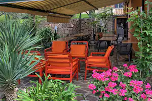 Orange Sitzgruppe auf der Camping-Terrasse unter Sonnenschirm, mit Steinterrasse, Steinmauer, Pflanzen und pinken Blüten im Garten.