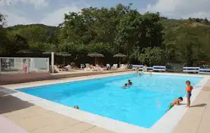 Schwimmbecken auf dem Campingplatz mit Liegen, Sonnenschirmen und Bäumen im Hintergrund, daneben ein Bereich mit weiteren Sitzplätzen.