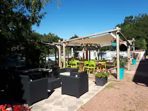Terrasse mit Pergola und grünen Gartenstühlen, Pflanzkübeln und Steintisch, im Hintergrund Bäume und ein Weg.