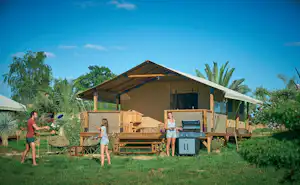 Camping-Unterkunft mit Holzplattform und Vorzelt, daneben Grill und Sitzbank; im Hintergrund Palmen und grünes Gelände.