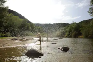 Fluss mit flachen Steinen und zwei Wasserschildkröten, umgeben von grünen Ufern und bewaldeten Hügeln.