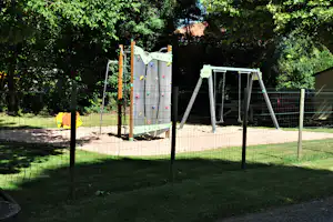 Spielplatz mit Kletterwand und Schaukeln im Sand, umgeben von Zäunen und viel Grün im Hintergrund.