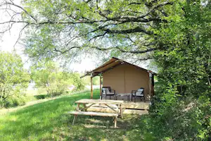 Holztisch und Sitzgruppe vor einem Zeltunterstand auf einer Campingwiese, umgeben von Bäumen und grünem Gras.