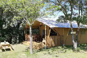 Campingzelt mit Holzpodest und Sitzbank im Grünen, umgeben von Bäumen und Rasenfläche.