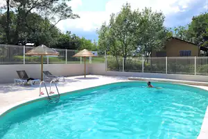 Camping-Pool mit türkisfarbenem Wasser, zwei Liegen und Sonnenschirmen, dahinter Zaun, Bäume und ein Gebäude.