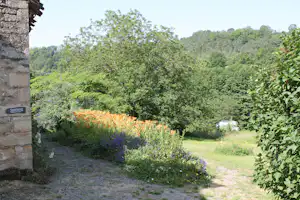 Blick auf einen Campinggarten mit Blumenbeeten, Rasen und Bäumen, links ein Steingebäude mit Schild „Sanitaire“.