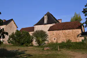 Stein Gebäude mit rotem Ziegeldach, Rasenfläche und Büschen im Vordergrund, dahinter weitere Häuser unter blauem Himmel.