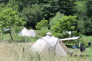 Campingzelt auf einer Wiese zwischen Bäumen und Sträuchern, im Hintergrund weitere Zelte und ein Sonnenschirm.