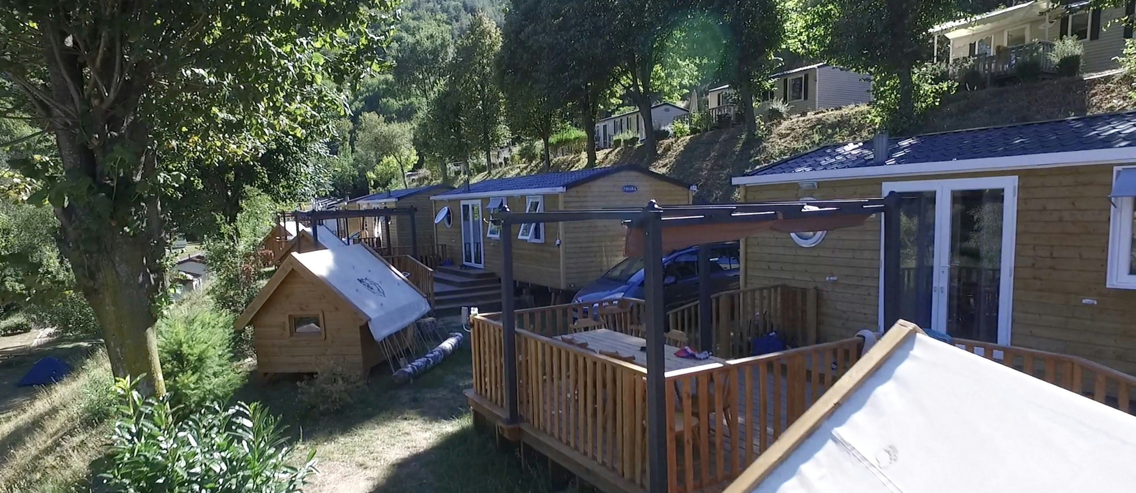 Reihe von Bungalows mit Holzterrassen und großen Fenstern, im Hintergrund weitere Unterkünfte zwischen Bäumen und Grün.