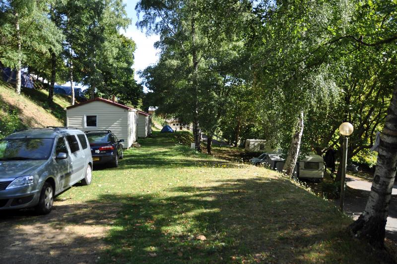 Grüner Campingplatz mit Rasenfläche, zwei Autos und einem kleinen Gebäude am Rand, umgeben von Bäumen.
