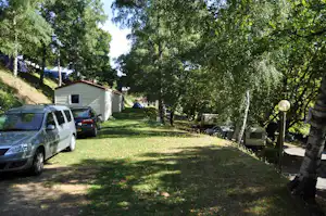 Grüner Campingplatz mit Rasenfläche, zwei Autos und einem kleinen Gebäude am Rand, umgeben von Bäumen.