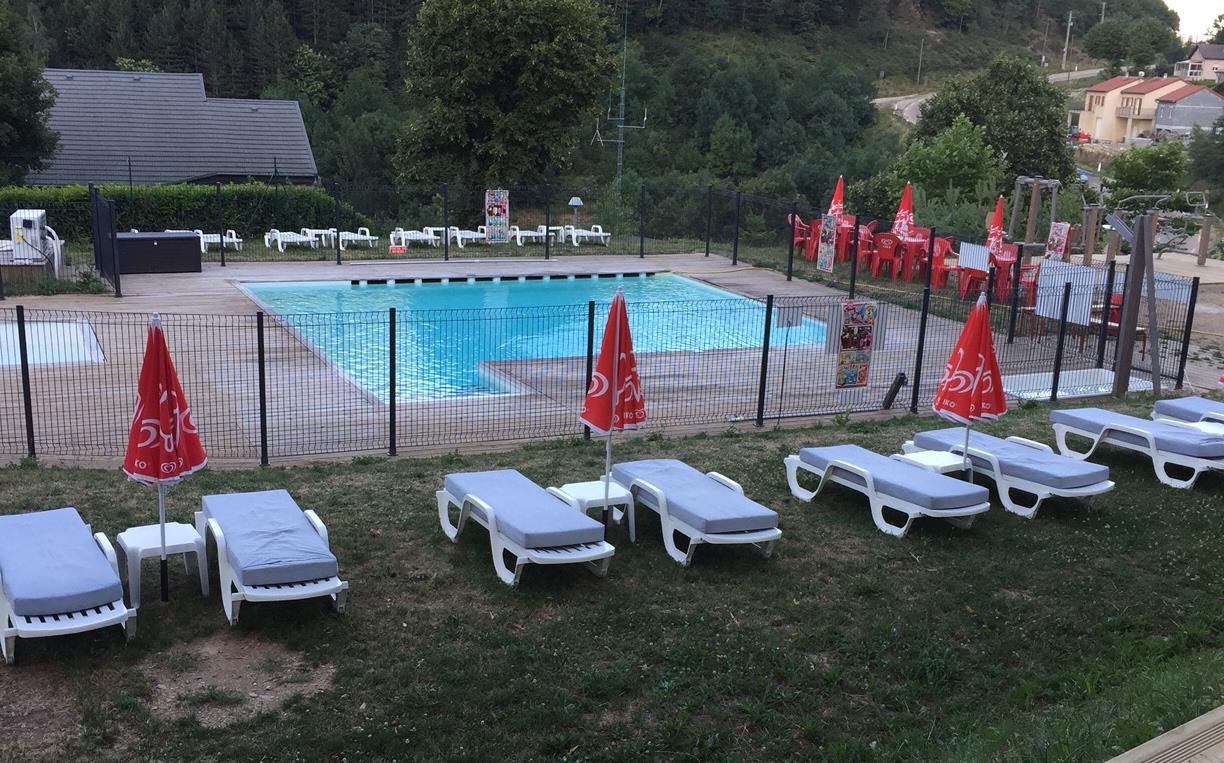 Schwimmbecken mit blauem Wasser, umzäunt mit roten Fahnen, Liegen auf dem Rasen und Bungalows im Hintergrund.