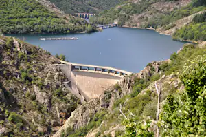 Großer Stausee mit Staumauer im engen Tal, umgeben von grünen Hügeln und Felswänden.