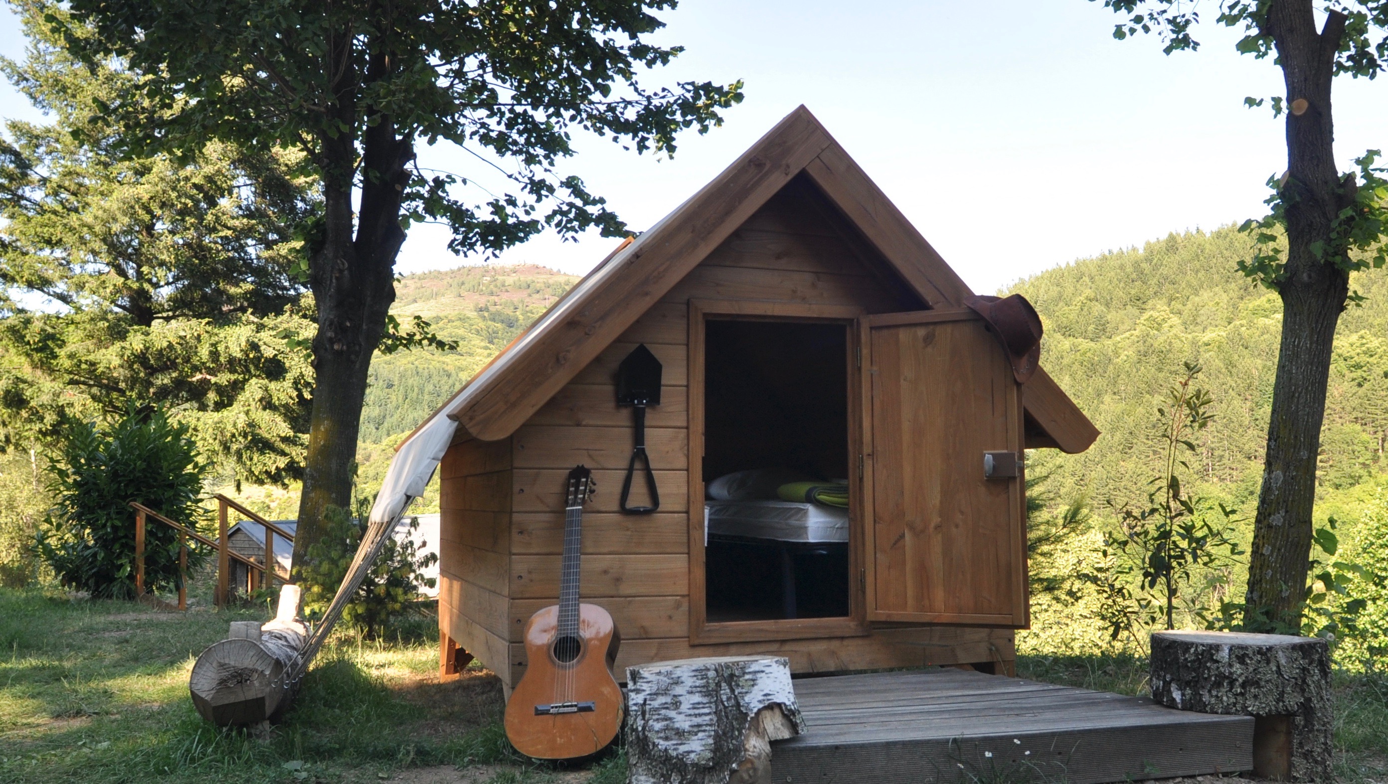 Holzsauna mit geöffnetem Eingang, davor Bank und Tisch, umgeben von Bäumen und grünen Hügeln im Hintergrund.