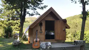Holzsauna mit geöffnetem Eingang, davor Bank und Tisch, umgeben von Bäumen und grünen Hügeln im Hintergrund.