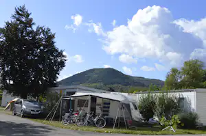 Wohnwagen mit Vorzelt und Fahrrädern auf dem Stellplatz, im Hintergrund Bäume und ein bewaldeter Hügel unter blauem Himmel.