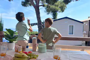 Zwei Personen sitzen am Camping-Tisch mit Getränken und Obst, im Hintergrund Wohnwagen und Bungalows, Bäume und Himmel.