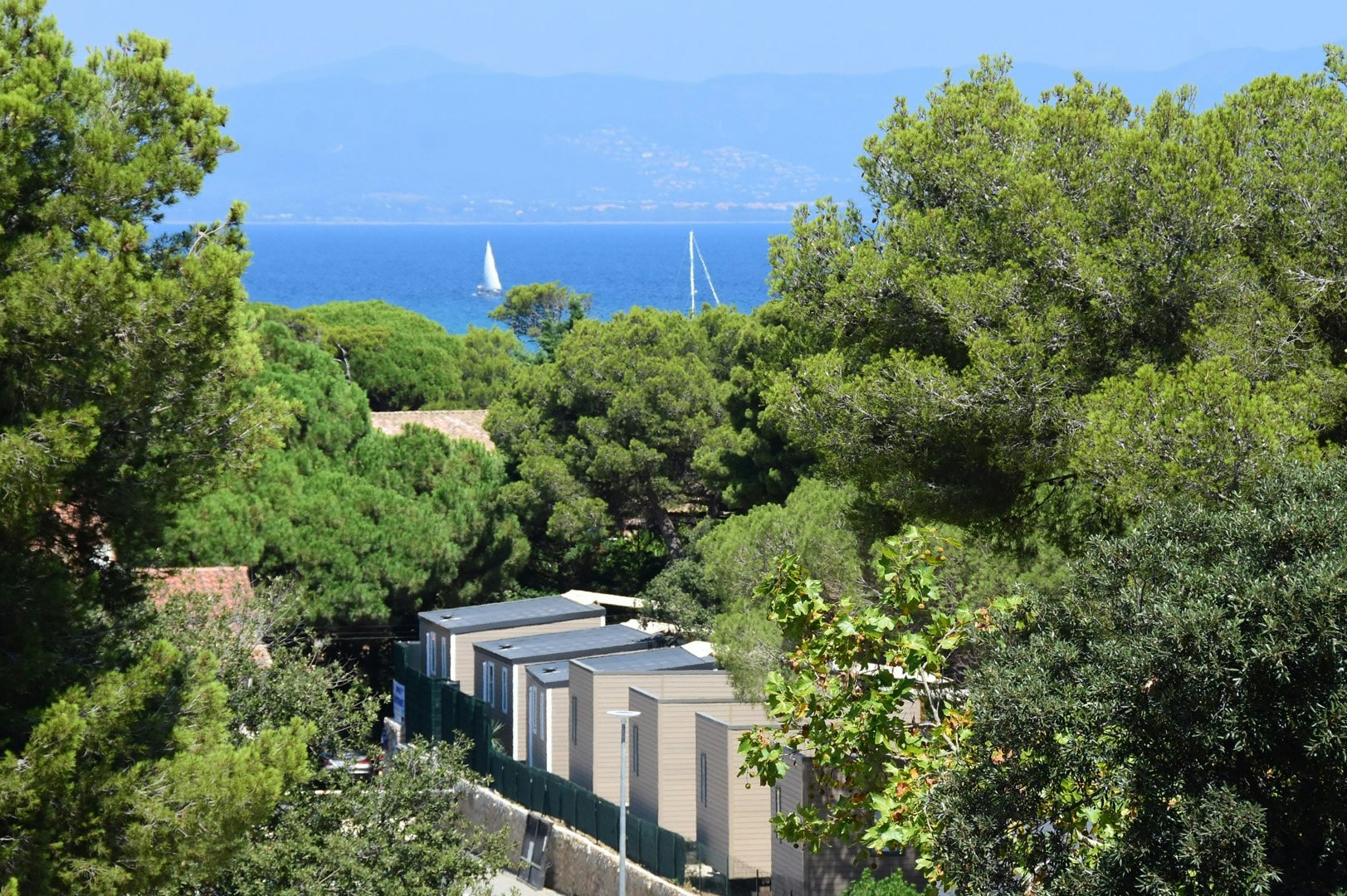 Blick auf moderne Campingunterkuenfte zwischen Gruenpflanzen, dahinter das Meer und Segelboote vor den Bergen.