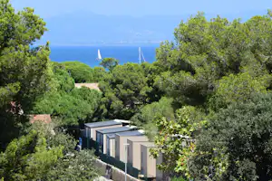 Blick auf moderne Campingunterkuenfte zwischen Gruenpflanzen, dahinter das Meer und Segelboote vor den Bergen.
