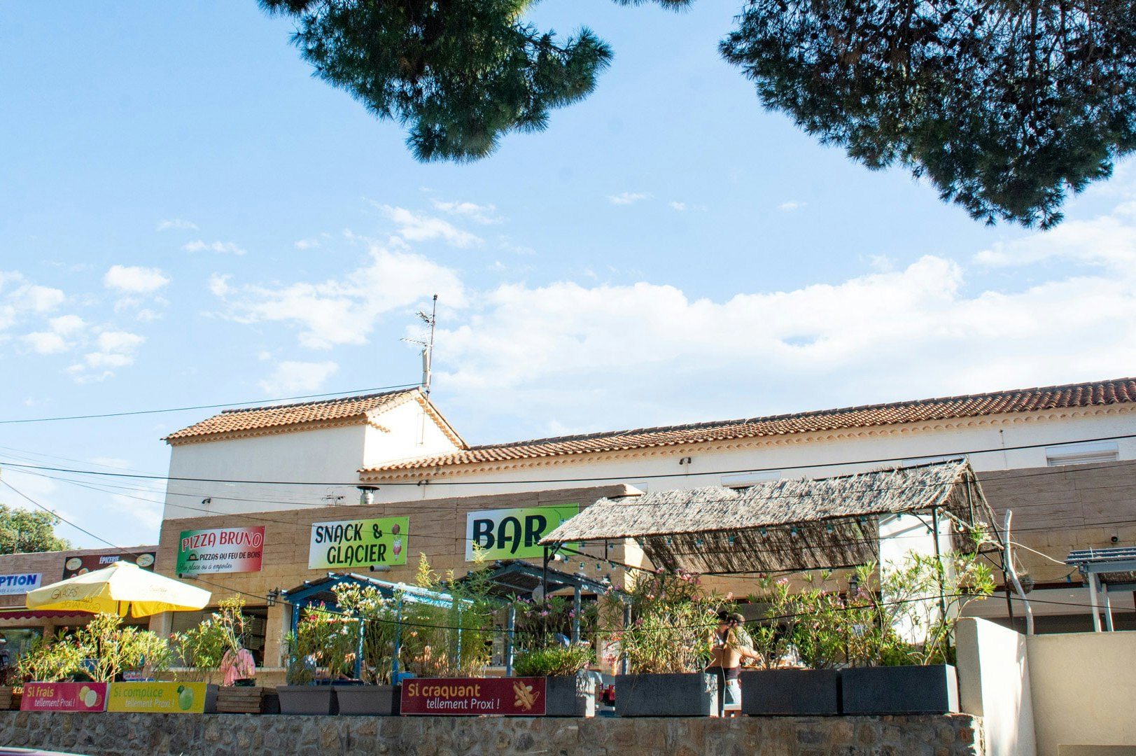 Überdachte Terrasse eines Campingplatz-Restaurants mit Sonnenschirmen, Gebäudefassade und Schildern im Hintergrund.