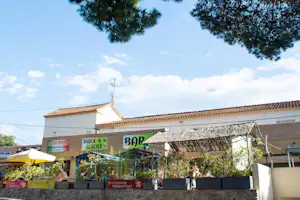 Überdachte Terrasse eines Campingplatz-Restaurants mit Sonnenschirmen, Gebäudefassade und Schildern im Hintergrund.