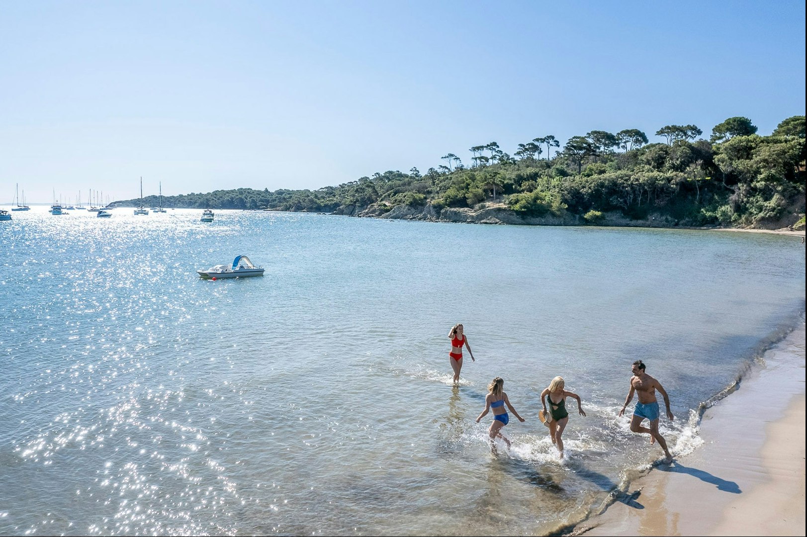 Badegäste im flachen Wasser am Strand, im Hintergrund Segelboote und eine bewaldete Küste.