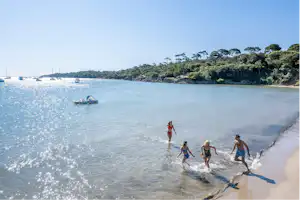 Badegäste im flachen Wasser am Strand, im Hintergrund Segelboote und eine bewaldete Küste.