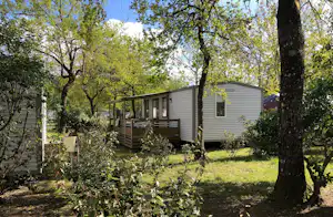 Mobilheim mit Holzterrasse im Grünen, umgeben von Bäumen und Sträuchern, im Campingplatzbereich.