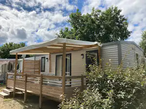 Mobilheim mit Holzveranda und Geländer, davor Sträucher und Bäume, im Hintergrund ein weiteres Gebäude am Campingplatz.