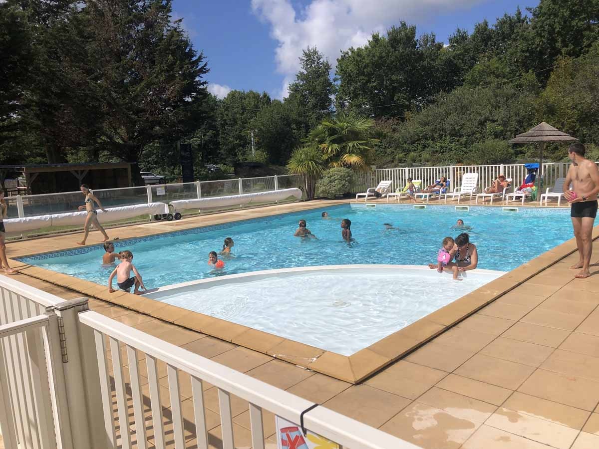 Camping-Pool mit Wasserfläche, weißen Liegen am Beckenrand und Zaun, im Hintergrund Bäume und ein Sonnenschirm.
