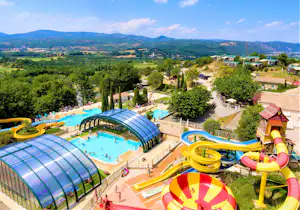 Beheiztes Freibad mit überdachten Becken, bunten Wasserrutschen und Blick auf grüne Hügel und Berge.