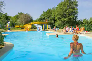 Schwimmbecken auf dem Campingplatz mit Rutsche, aufblasbarem Spielbereich und Kindern im Wasser vor Bäumen und Sonnenschirmen.