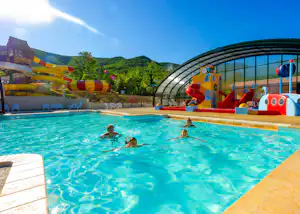 Schwimmbecken auf dem Campingplatz mit Wasserfontänen, aufblasbarem Spielbereich und Rutsche, im Hintergrund Bäume und Hügel.