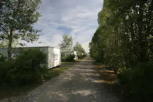 Campingweg zwischen Bäumen und Gebäuden, links ein langes Steinwand-Gebäude, rechts Hecken und ein weiterer Wegabschnitt.