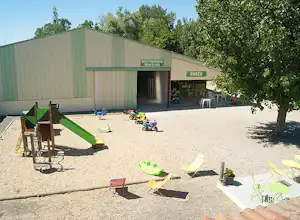 Campingplatz mit gepflastertem Hof, Sitzgelegenheiten und Stühlen vor einem Gebäude, im Hintergrund Bäume und ein Fahrrad.