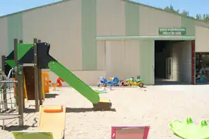 Spielplatz am Campingplatz: grüne Rutsche, Sandfläche und Spielzeug vor einer großen Halle mit Eingang.